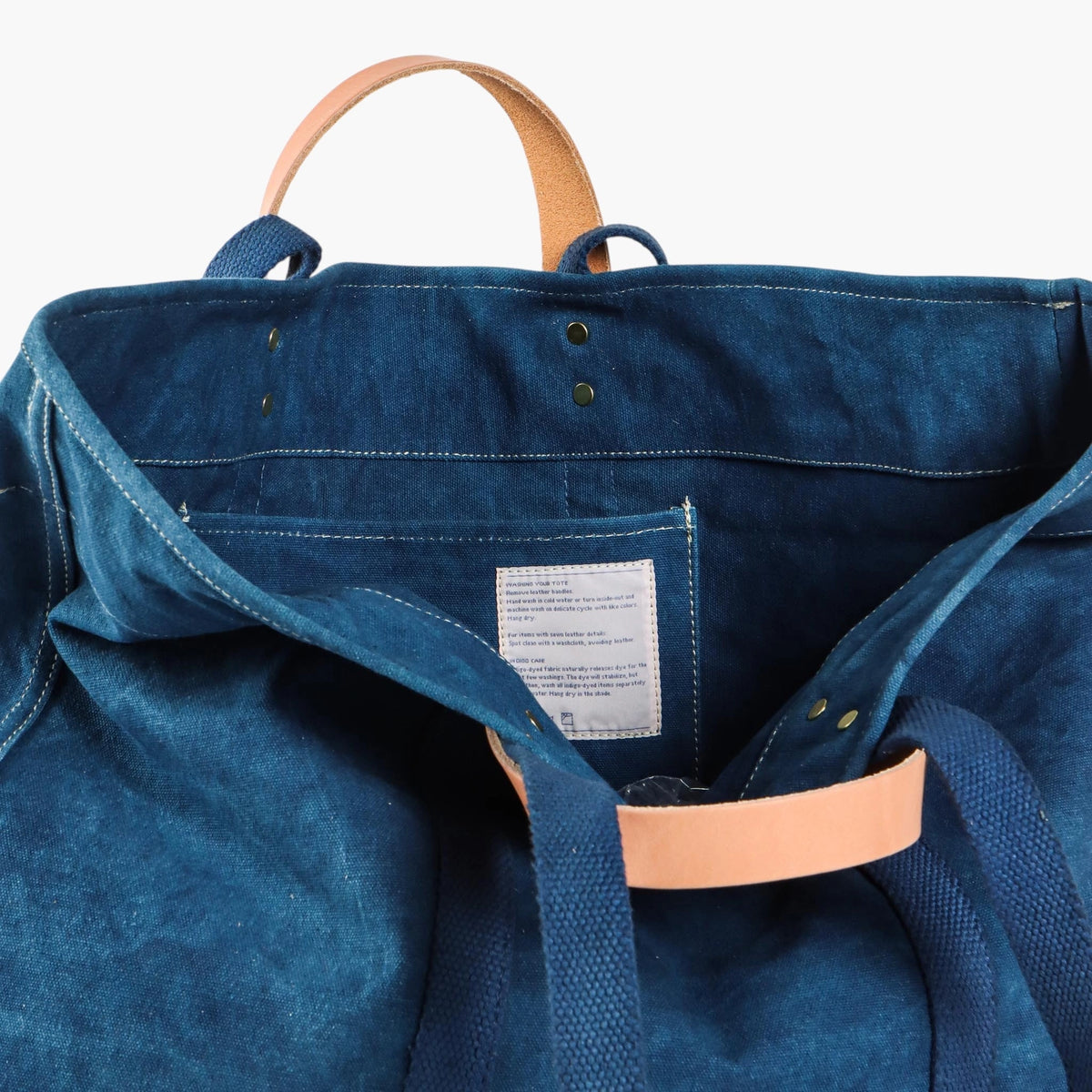 Interior of the indigo canvas tote showing the pocket, care label, and leather and webbing handles.
