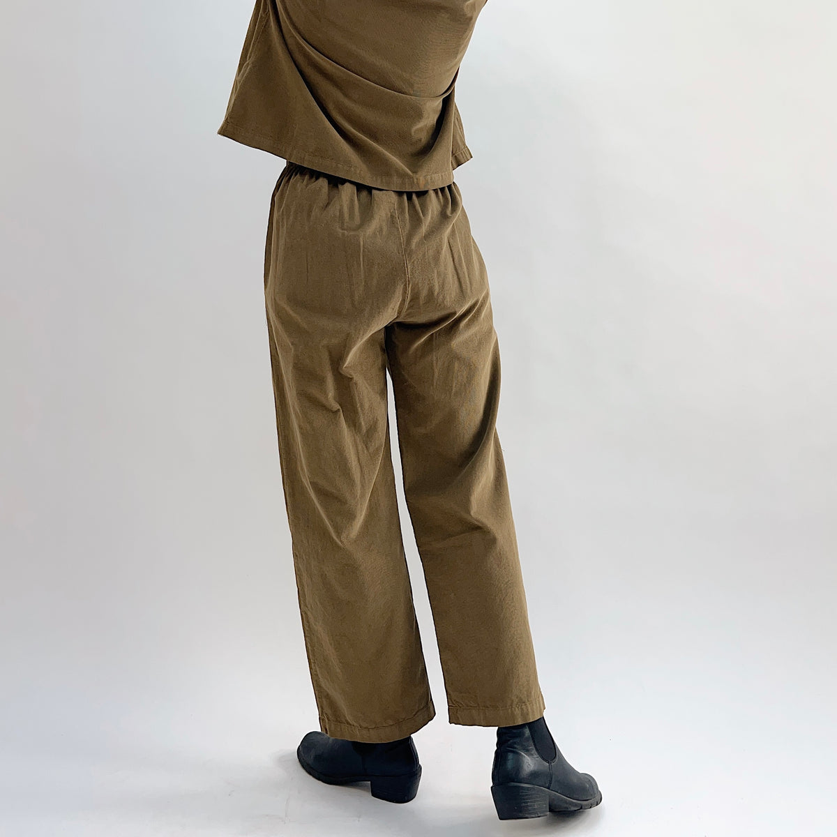 Back and side view of the Bryn Walker Corduroy Pleated Pants in Partridge, showing elastic back waistband, straight leg, full-length hem, and olive pinwheel corduroy drape, styled with matching jacket and black Chelsea boots
