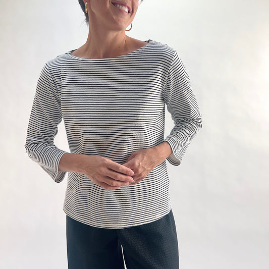 Smiling front view of model wearing a white and black horizontal stripe boatneck tee with 3/4 sleeves, hands clasped at waist, showing the wide boatneck neckline and crinkle-texture fabric, styled with black wide-leg pants against a white background.
