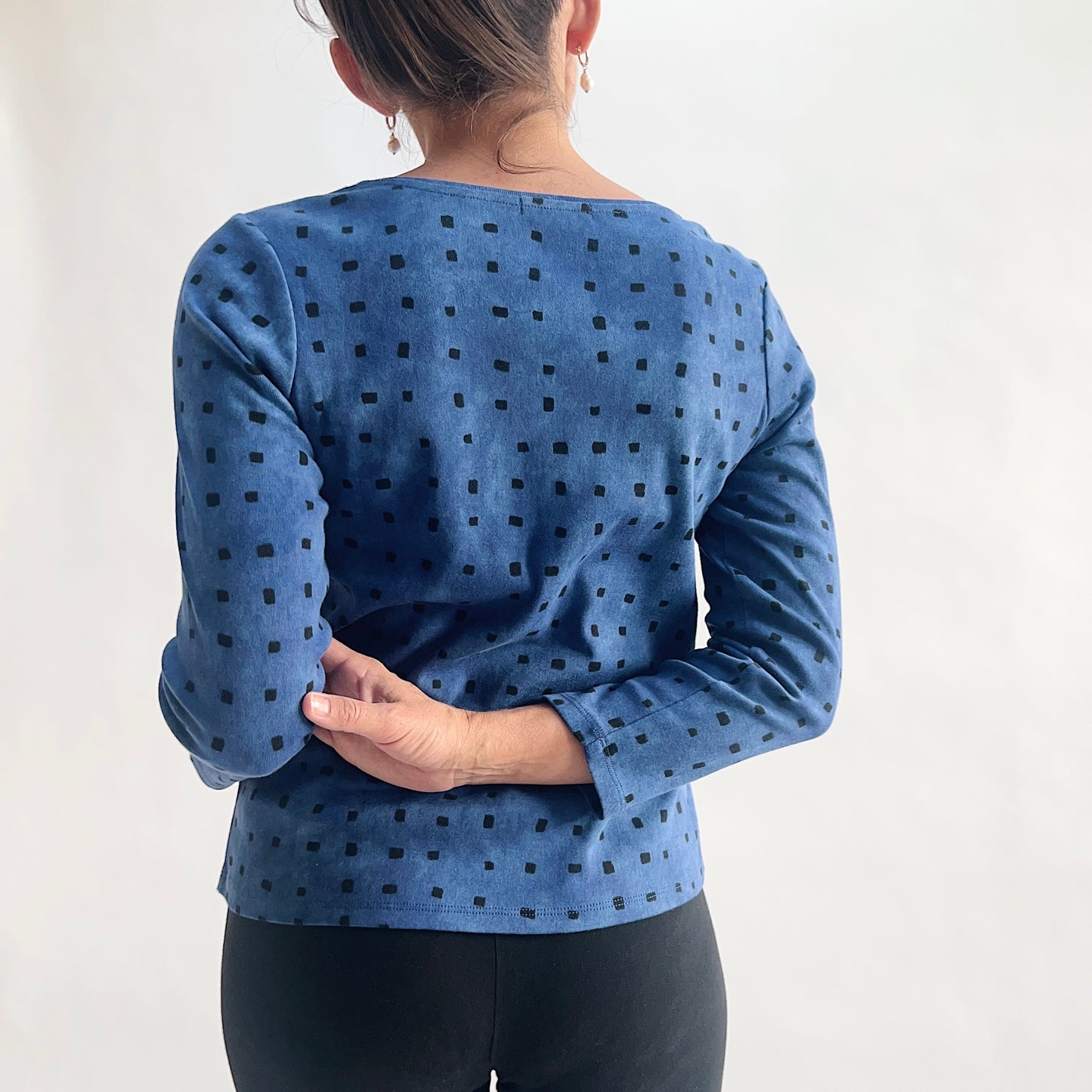 Back view of model wearing a medium indigo long-sleeve cozy rib tee with all-over tonal dark navy square print, arms clasped behind back, showing the scoop back neckline and relaxed fit, styled with black pants against a white background.
