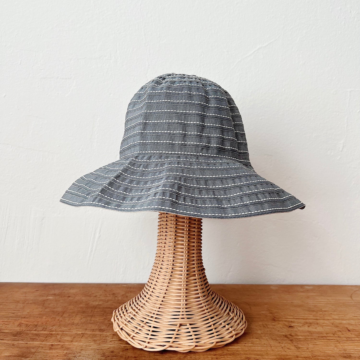 Sage ribbon bucket hat with white stitch stripe detail and wide brim, shown on a wicker hat stand — front view