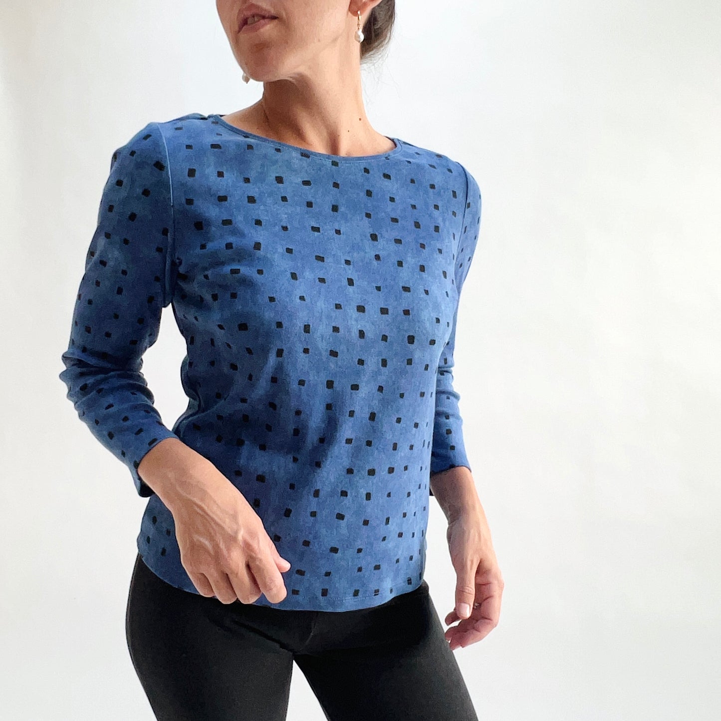 Three-quarter front view of model wearing a medium indigo long-sleeve cozy rib tee with all-over tonal dark navy square print, arms in motion, showing the relaxed fit and hem, styled with black pants against a white background.
