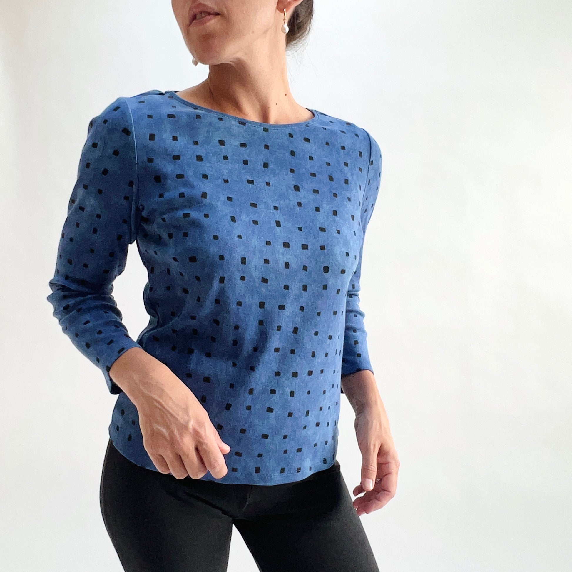 Three-quarter front view of model wearing a medium indigo long-sleeve cozy rib tee with all-over tonal dark navy square print, arms in motion, showing the relaxed fit and hem, styled with black pants against a white background.
