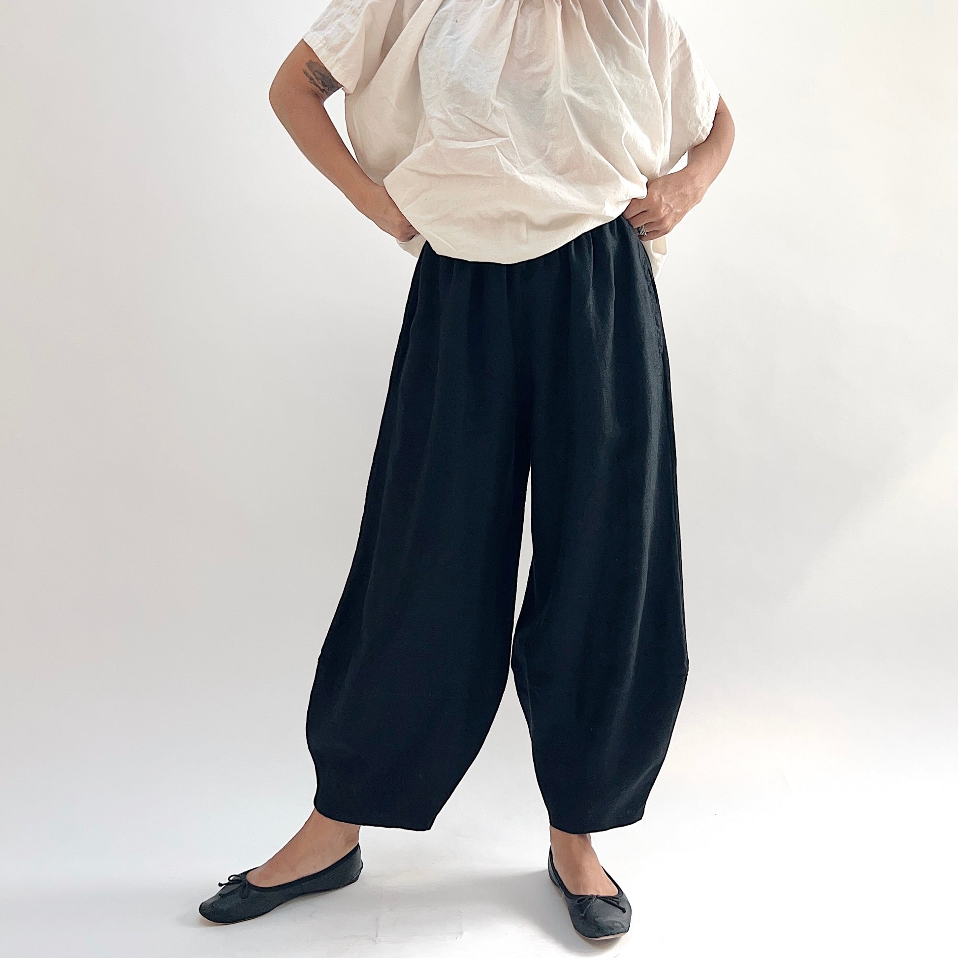 Model with hands on hips wearing the Bryn Walker Heavy Linen Oliver Pant in Black, full front view showing barrel-leg silhouette, elastic waistband, and tapered ankle in black heavy linen, styled with cream gathered linen top and black ballet flats
