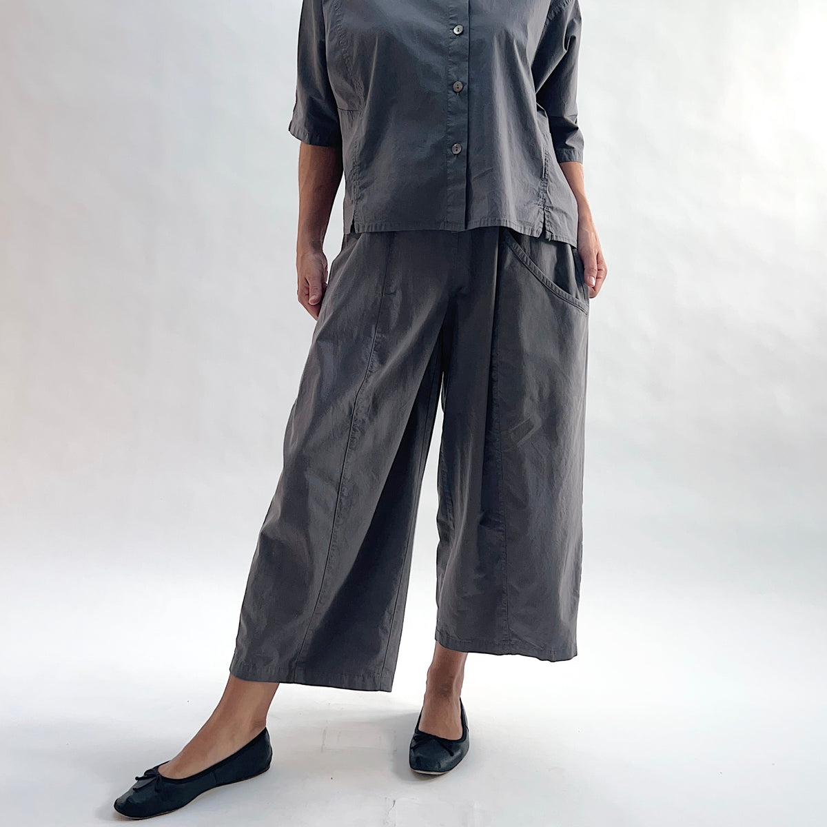 Full front view of model wearing Eleven Stitch eiffel tower one pocket pant with hands in pockets, showing elastic waistband, wide curved-seam cropped leg, large side pocket, gathered elastic hem, and deep slate grey color, styled with matching grey short sleeve button front shirt and black ballet flats