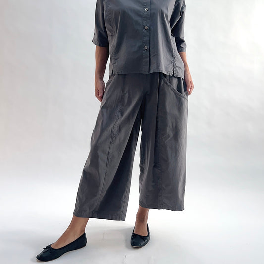 Full front view of model wearing Eleven Stitch eiffel tower one pocket pant with hands in pockets, showing elastic waistband, wide curved-seam cropped leg, large side pocket, gathered elastic hem, and deep slate grey color, styled with matching grey short sleeve button front shirt and black ballet flats