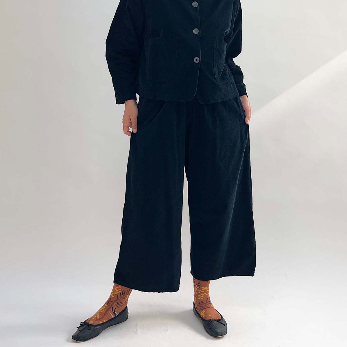 Model wearing the Bryn Walker Corduroy Flood Pants in Black, full front view showing elastic waistband, relaxed wide-leg flood silhouette, and black pinwheel corduroy, paired with matching Standing Collar Crop Jacket, printed socks, and black ballet flats
