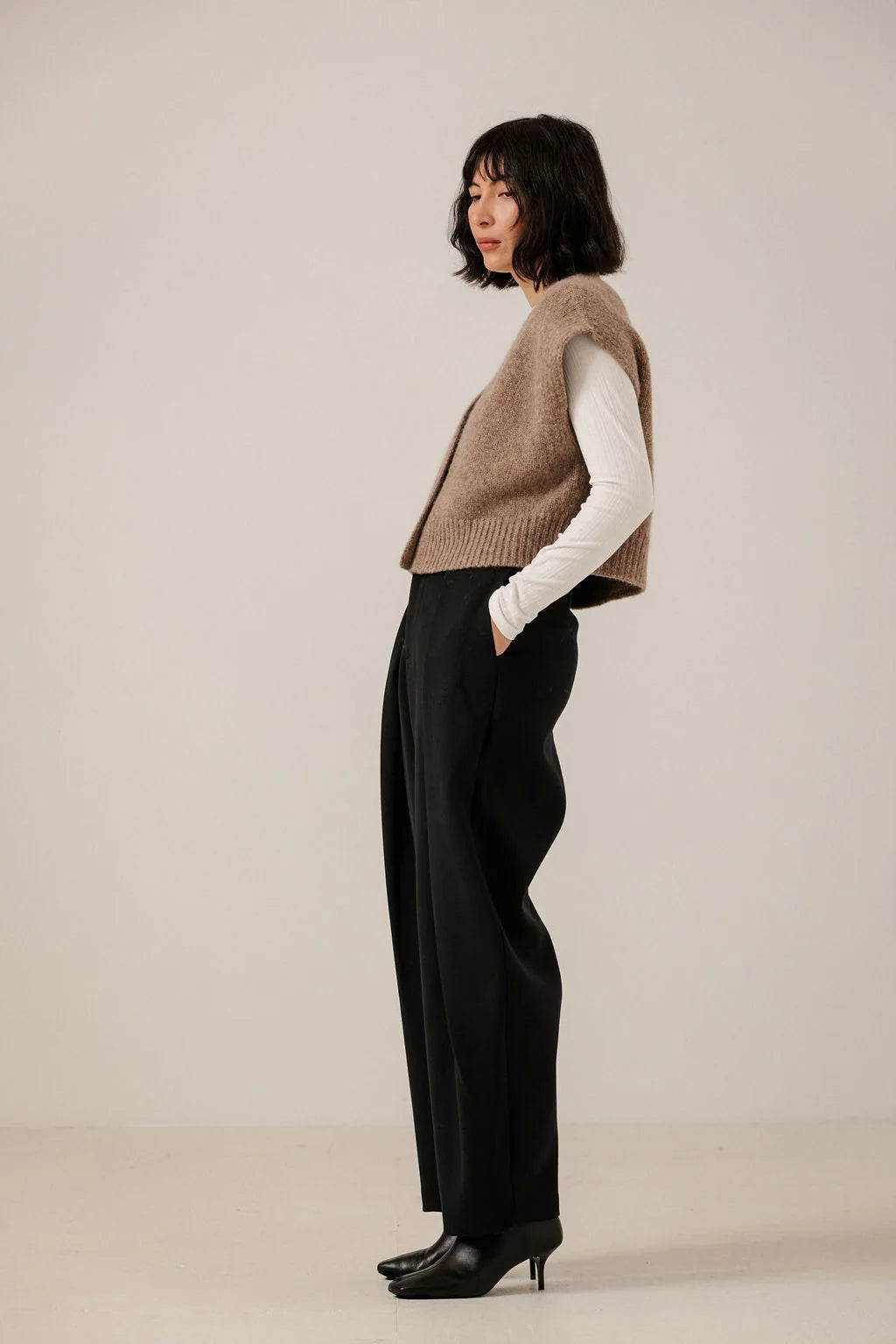 Side view of a model wearing the Dark Fawn vest over a white long-sleeve top with black trousers.
