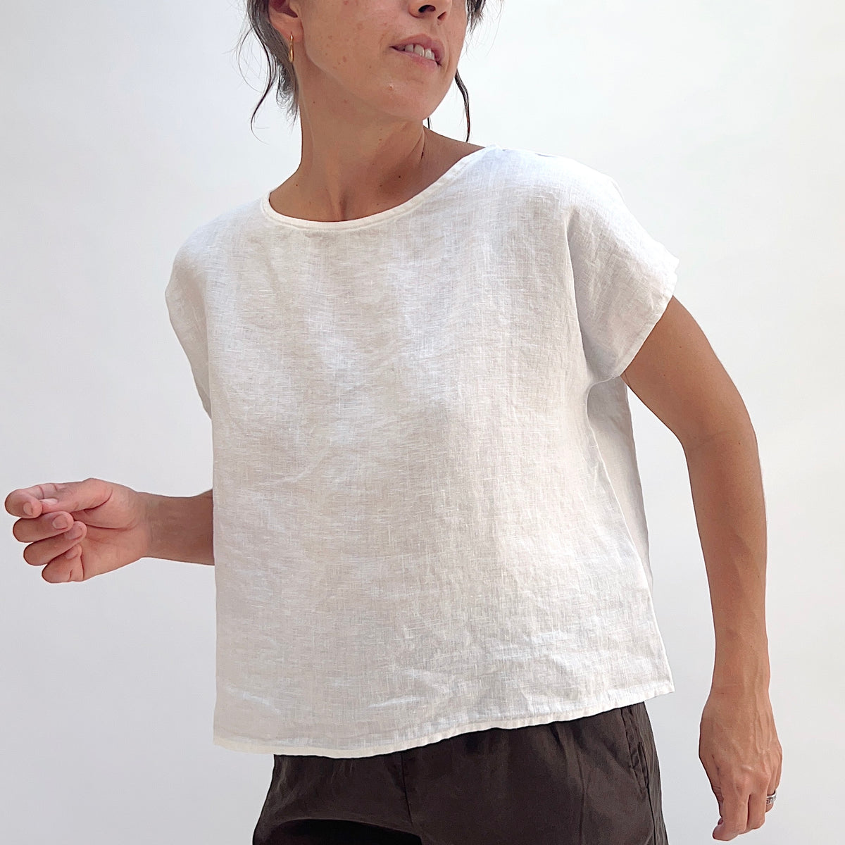 Close-up front view of white linen high-low tee highlighting boatneck neckline, cap sleeves, and woven linen texture, styled with dark trousers
