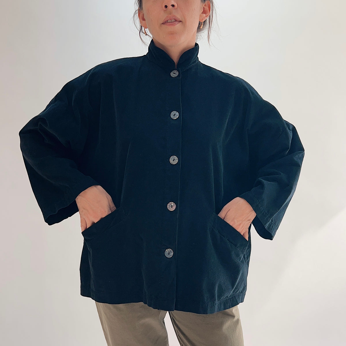Model with both hands in pockets wearing the Bryn Walker Corduroy Danuta Jacket in Black, full front view highlighting standing collar, shell button closure, wide dolman sleeves, two front patch pockets, and relaxed oversized fit in black pinwheel corduroy, styled with olive pants
