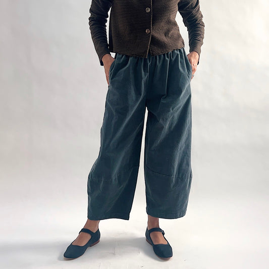 Front view of model wearing iron blue-gray mini cord lantern pants with hands in side pockets, showing the gathered elastic waist, wide lantern silhouette, and tapered ankle, styled with the matching chocolate striped cropped cardigan and blue mary jane flats against a white background.
