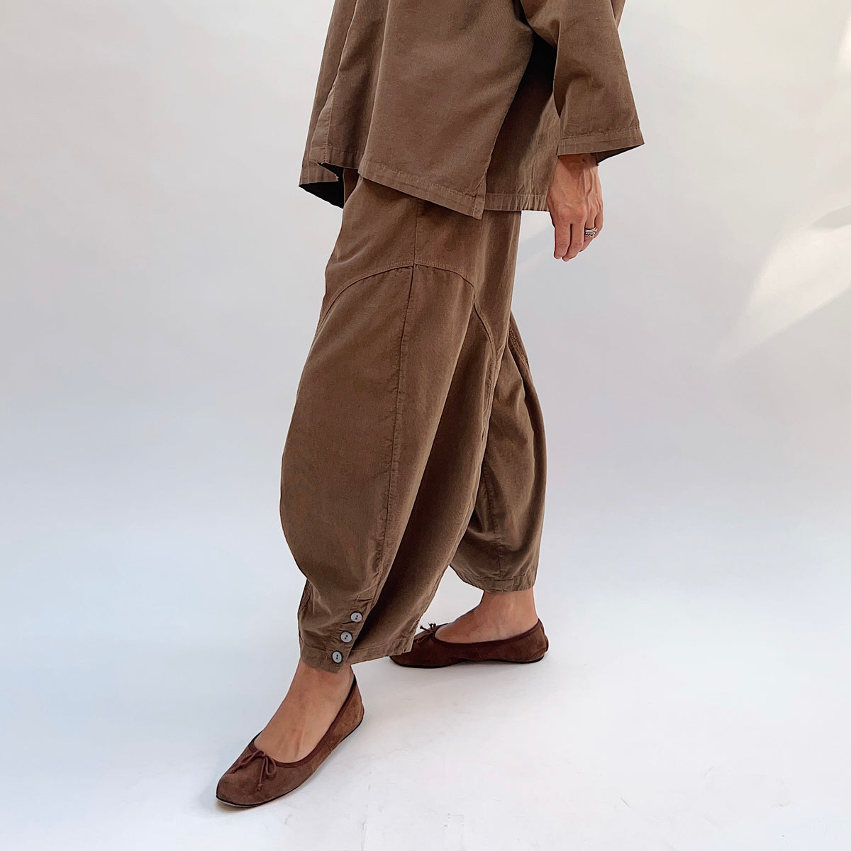 Eleven Stitch Corduroy Doreen Pant in Winter Olive, angled side view highlighting the curved seam detail on the balloon leg and three-button gathered ankle cuff, styled with a matching corduroy top and brown ballet flats
