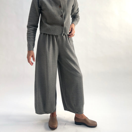 Full front view of model wearing squirrel and black fine horizontal stripe wide-leg cropped pants, hands on hips showing the elastic waist and dart shaping, styled with the matching striped asymmetrical cardigan and taupe slip-on shoes against a white background.
