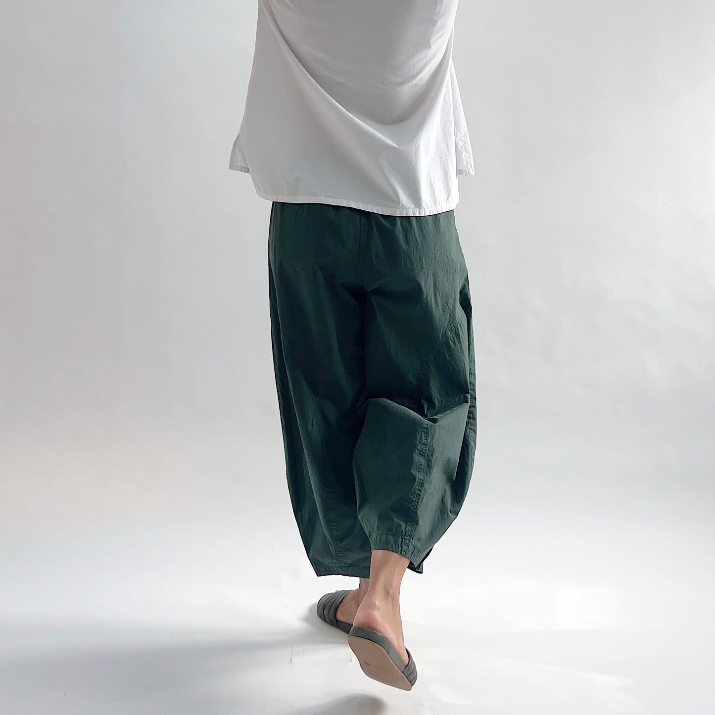 Three-quarter back view of model wearing Eleven Stitch konbu snap doreen pant in motion, showing elastic waistband, wide balloon leg tapering to snap hem at ankle, side seam detailing, snap buttons at ankle, and deep forest green color, styled with white top and dark slide sandals