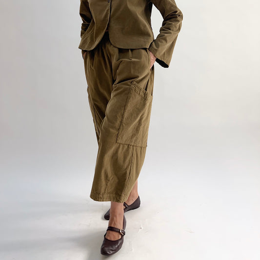 Model with hand on hip wearing the Bryn Walker Corduroy Pasha Pants in Partridge, front view showing side leg patch pockets, elastic waistband, and olive pinwheel corduroy, paired with matching Audrey Jacket and dark Mary Jane flats
