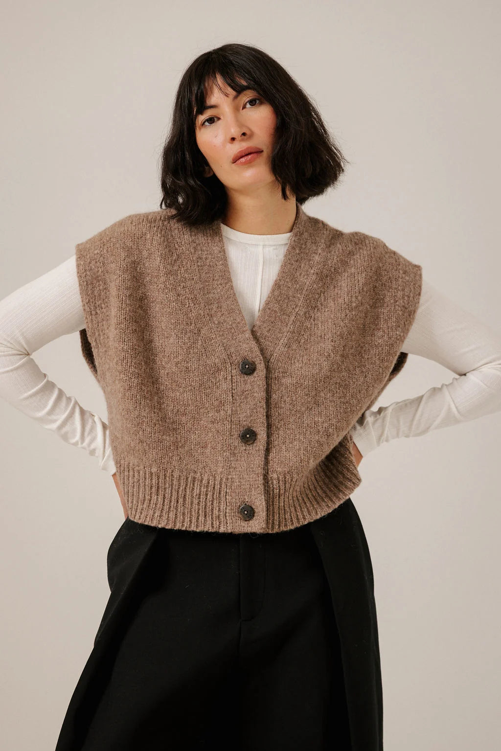 Front view of a model wearing the Dark Fawn knit vest over a white top, showing its V-neck, buttons and ribbed hem.
