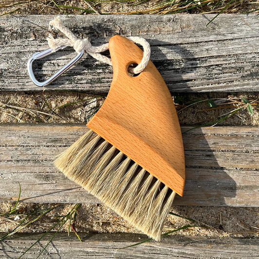A small hand brush with a light brown wooden handle, natural beige bristles, and a silver metal carabiner attached by a white rope, placed on weathered wooden planks outdoors.