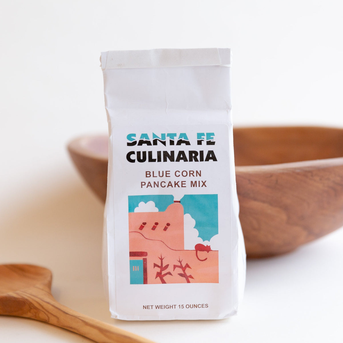 A white bag of Santa Fe Culinaria Blue Corn Pancake Mix stands upright on a light surface, with a wooden bowl and wooden spoon blurred in the background.