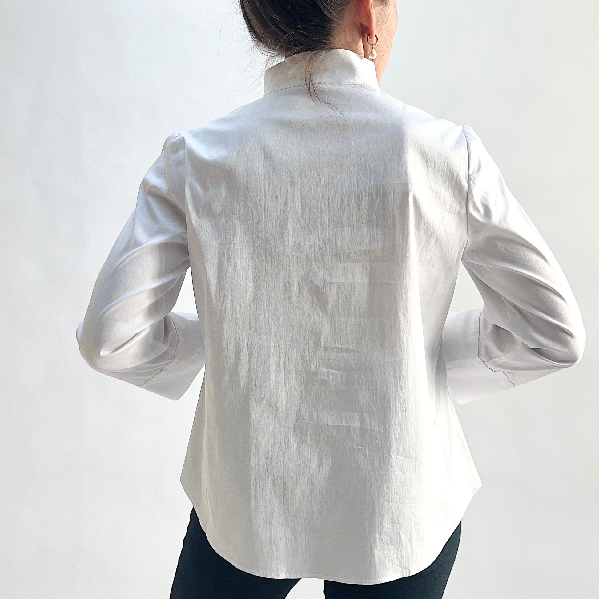 Back view of model wearing a white slubby cotton pullover shirt with standing band collar, 3/4 sleeves with wide cuffs, and curved shirttail hem, hands behind back, showing the clean back panel and slubby fabric texture, styled with black slim pants against a white background.

