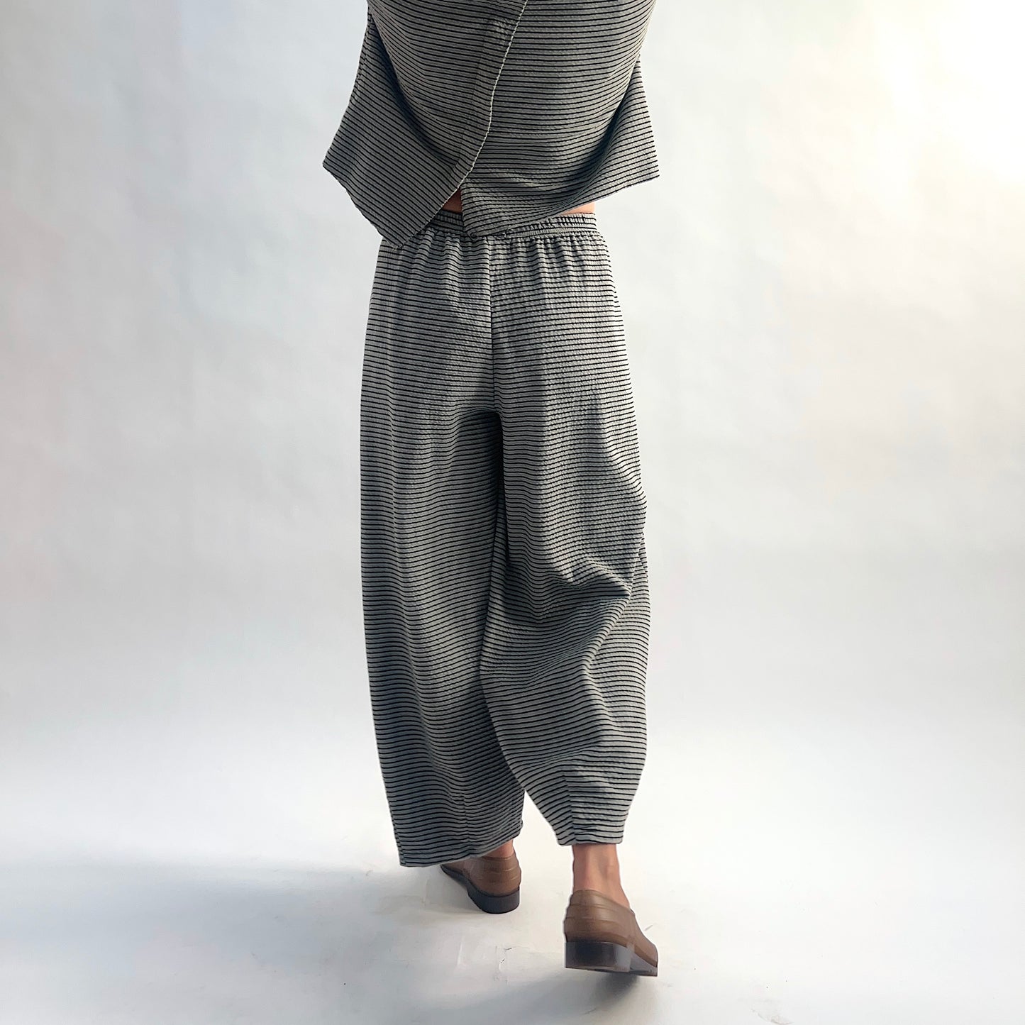 Back view of model wearing squirrel and black fine horizontal stripe wide-leg cropped pants with elastic waist, showing the relaxed wide-leg silhouette and crinkle-texture fabric, styled with the matching striped cardigan and taupe slip-on shoes.
