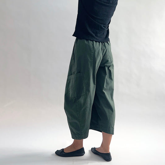 Back side view of model wearing Eleven Stitch konbu snap front pant, showing elastic back waistband, wide balloon leg, large patch pocket, gathered elastic hem, and deep forest green color, styled with black long sleeve top and black ballet flats