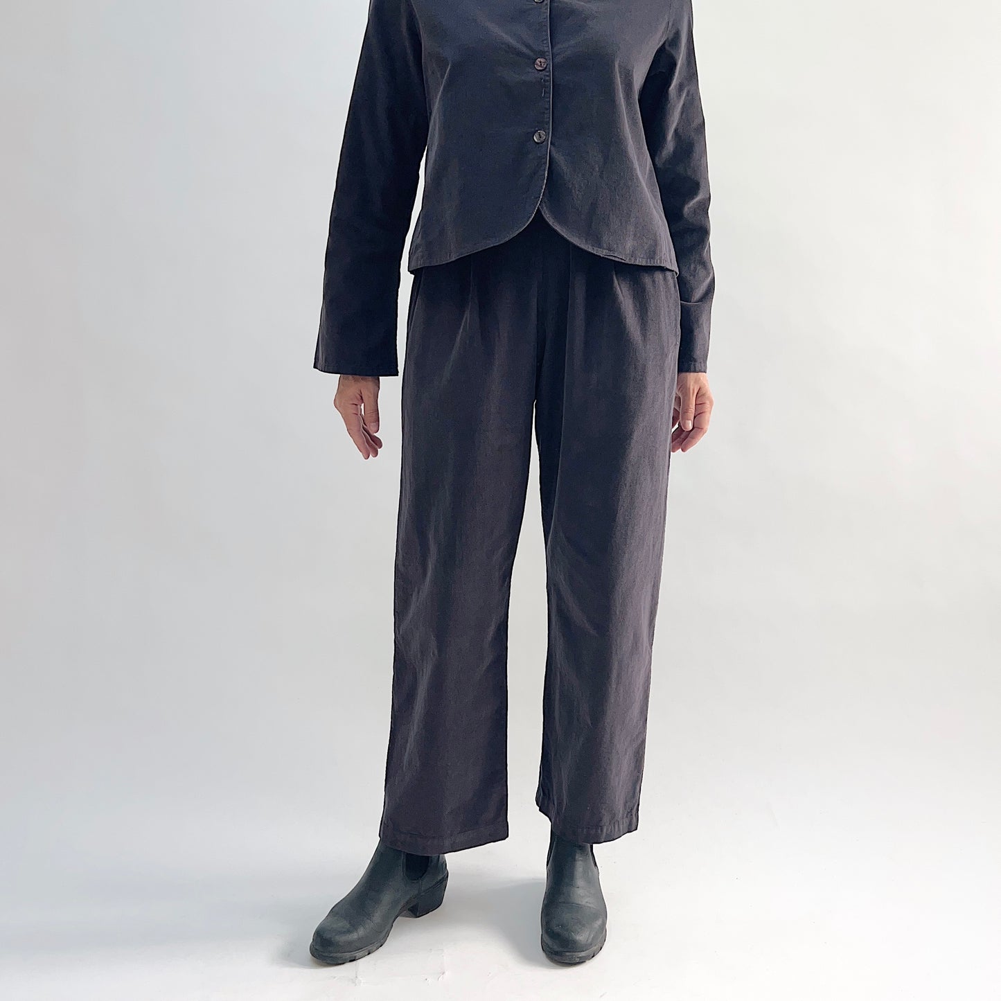 Model wearing the Bryn Walker Corduroy Pleated Pants in Fig, full front view showing flat front with pleats, elastic waistband, tapered straight leg, and deep fig pinwheel corduroy, styled with matching Standing Collar Crop Jacket and black Chelsea boots

