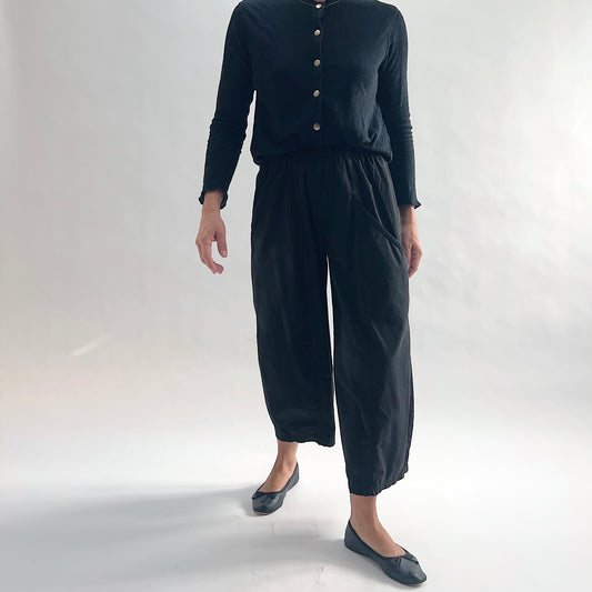 Three-quarter front view of model wearing Eleven Stitch black one pocket pant with arms at sides, showing elastic waistband, wide cropped leg, curved side pocket, gathered elastic hem, and black color, styled with black long sleeve button front top and black ballet flats