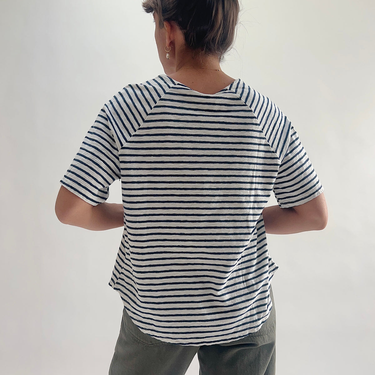 Back view of model wearing a navy and white striped boxy raglan tee, showing the curved hem and raglan sleeve seam detail, styled with olive green pants.
