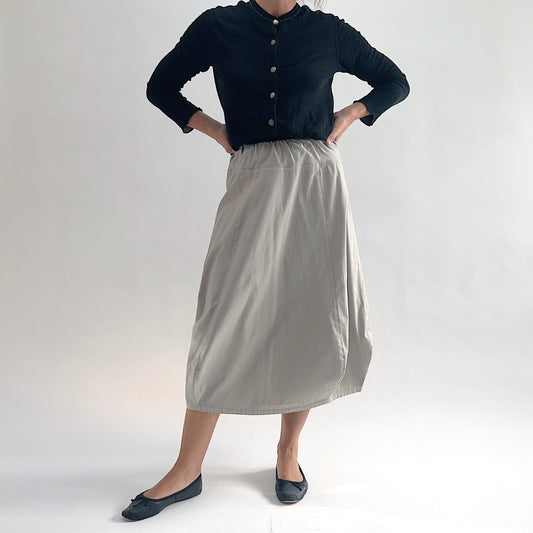 Three-quarter front view of model wearing Eleven Stitch fennel rib yoke skirt with hands at waist, showing elastic knit waistband, curved seam paneling, sculptural bubble hem, below-the-knee length, and warm taupe color, styled with black button-front cardigan and black ballet flats