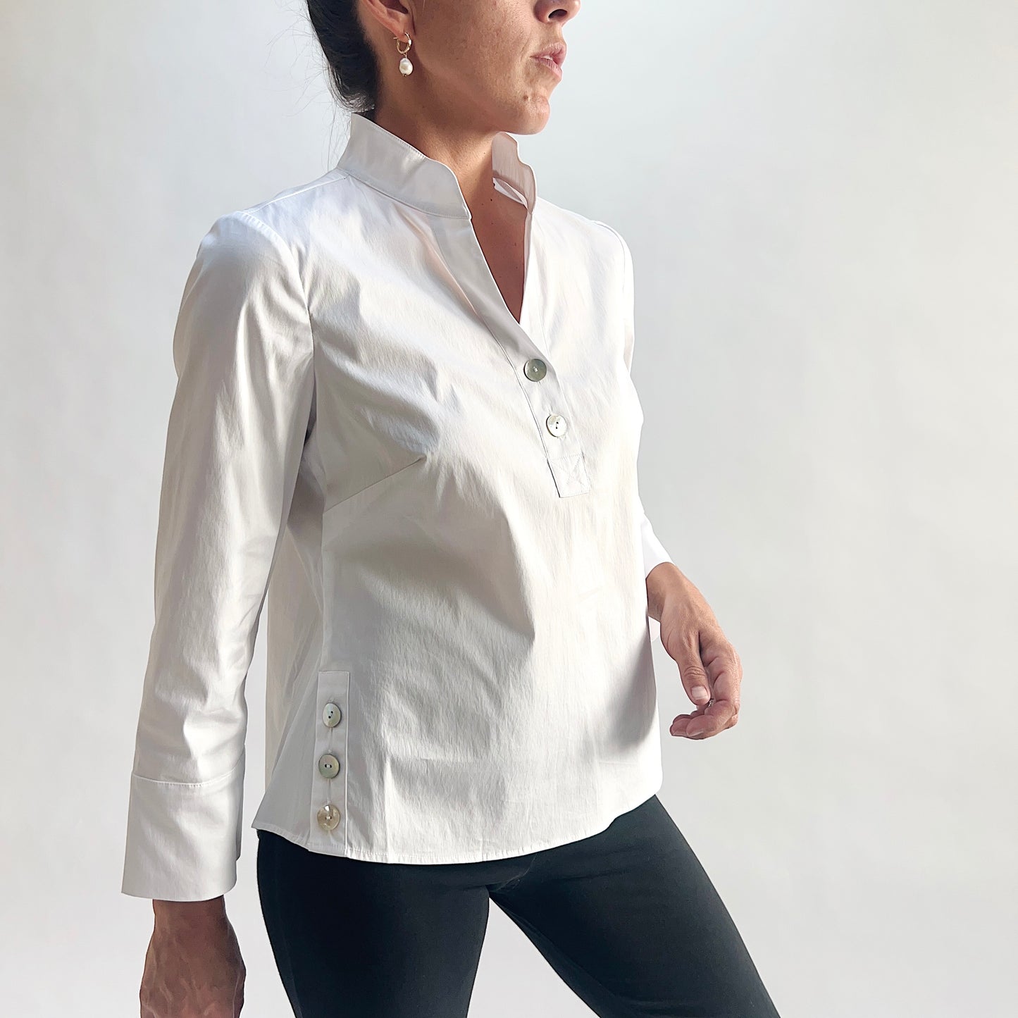 Three-quarter side view of model wearing a white slubby cotton pullover shirt, showing the standing band collar with V-notch, half placket with shell buttons, side hem button vents, and 3/4 sleeves with wide cuffs, styled with black slim pants against a white background.
