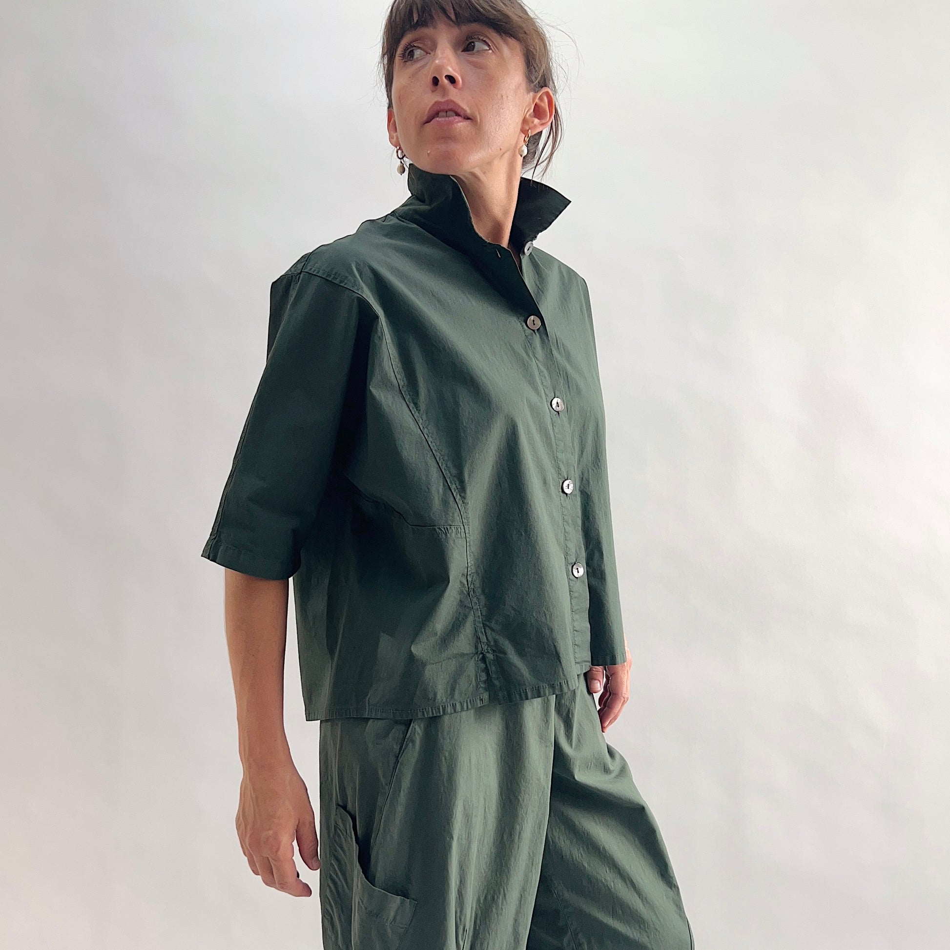 Three-quarter side view of model wearing Eleven Stitch konbu camp shirt looking up, showing stand collar, dark shell buttons, short sleeves, patch pocket, slightly cropped boxy fit, and deep forest green color, styled with matching konbu snap front pant