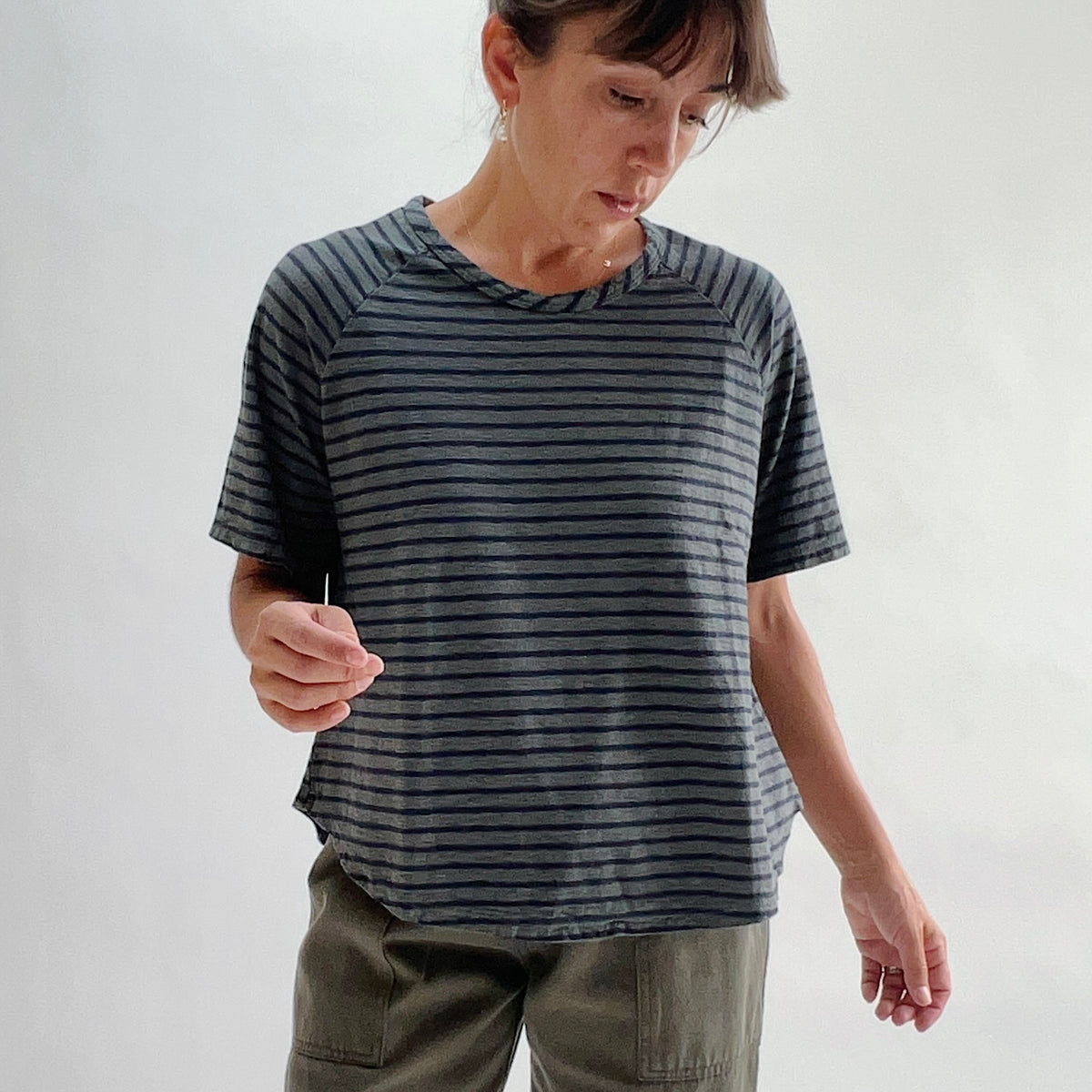Front view of model looking down wearing an anthracite and navy striped boxy raglan tee with a relaxed fit and boat neckline, paired with olive green pants.
