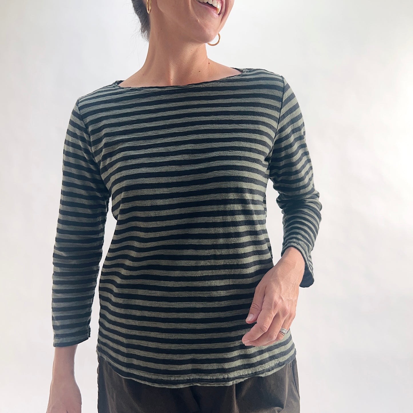 Smiling close front view of model wearing a kelp green and black horizontal stripe boatneck tee with 3/4 sleeves, hand on hip, showing the wide boatneck neckline and linen-blend texture, styled with dark charcoal pants.
