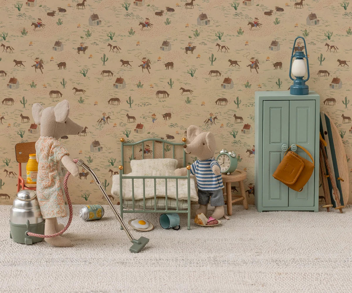 Maileg Vintage Single Bed in Mint — lifestyle scene of two Maileg mouse dolls in a playroom with western-themed wallpaper; a Big Sister mouse in a floral dress vacuuming near the mint single bed while a Little Brother mouse in a striped top holds an alarm clock beside a teal wardrobe
