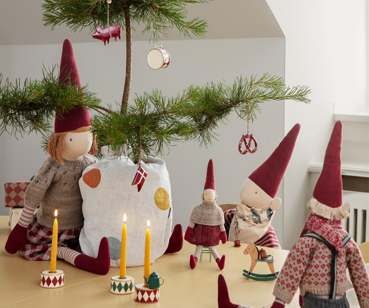 Maileg Pixy Girl in a knitted cape and red striped skirt, seated among larger pixy dolls beside a decorated Christmas tree and candles.
