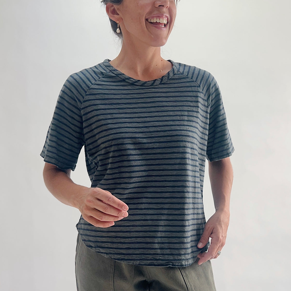 Close front view of model smiling in an anthracite and navy striped boxy raglan tee, showing the boat neckline and short raglan sleeves, styled with olive green pants.
