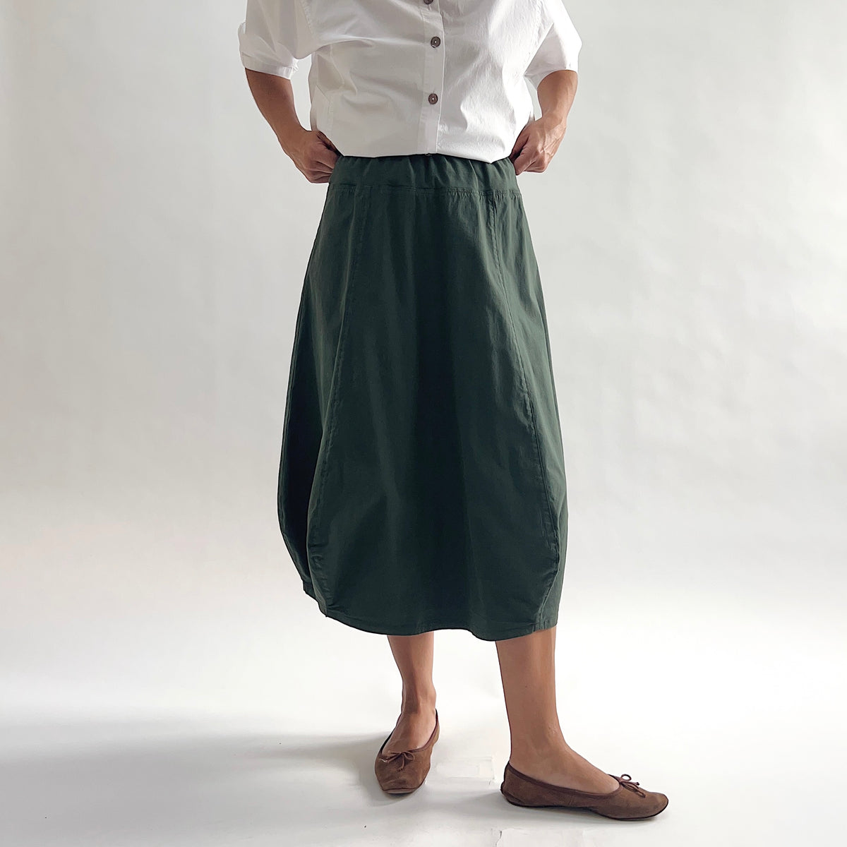 Full front view of model wearing Eleven Stitch konbu rib yoke skirt with hands on waist, showing elastic knit waistband, curved seam paneling, sculptural bubble hem, below-the-knee length, and deep forest green color, styled with white chalk camp shirt and brown ballet flats