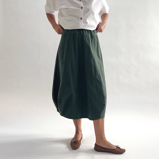 Full front view of model wearing Eleven Stitch konbu rib yoke skirt with hands on waist, showing elastic knit waistband, curved seam paneling, sculptural bubble hem, below-the-knee length, and deep forest green color, styled with white chalk camp shirt and brown ballet flats