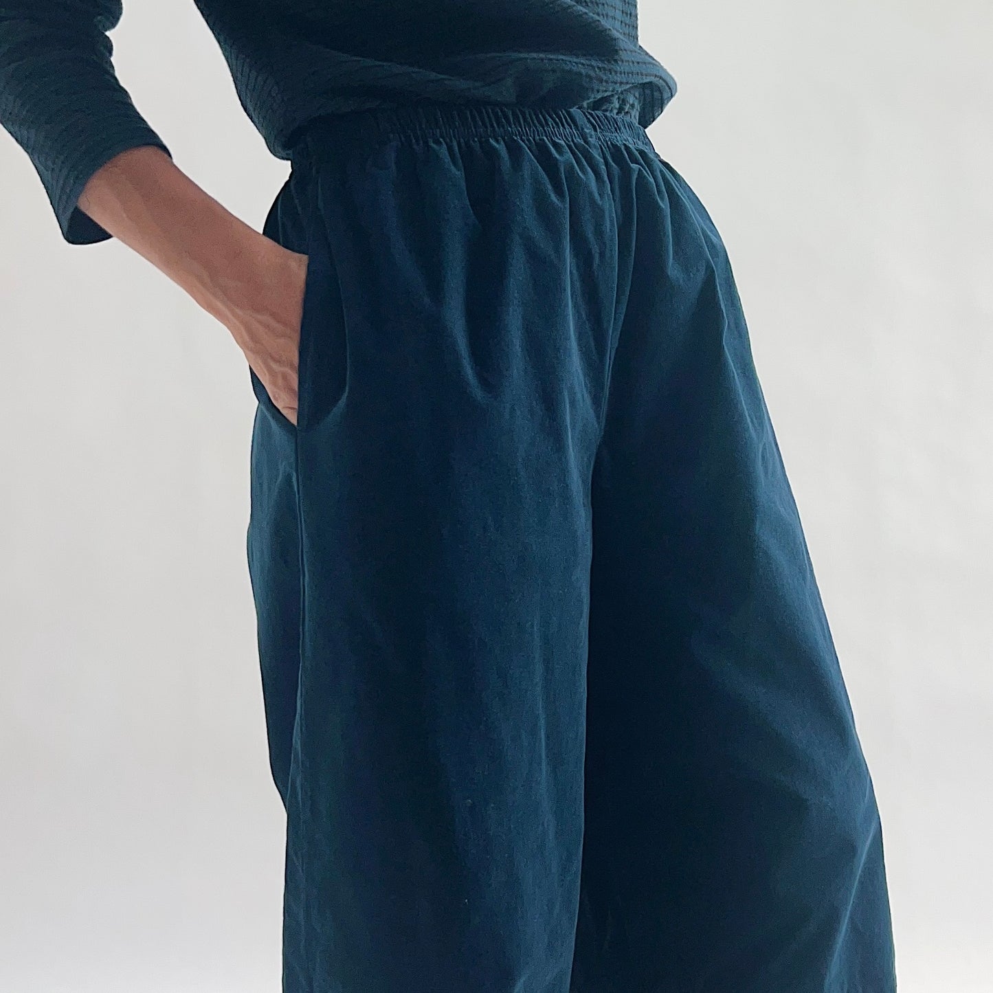 Close side view of model wearing deep teal mini cord lantern pants, hand in side pocket, showing the gathered elastic waist, fine cord texture, and wide lantern volume at the hip, styled with a matching teal long-sleeve top against a white background.
