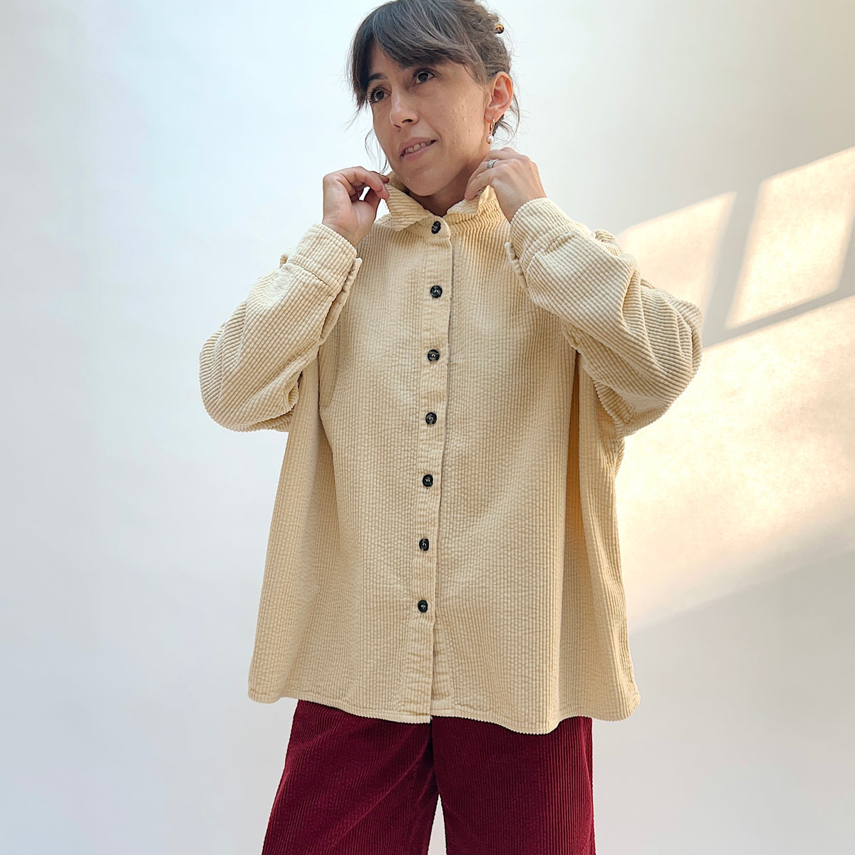 Model adjusting the collar of the Cut Loose Lux Corduroy Oversized Shirt in Glow, showing the pale cream fabric and dark front buttons.
