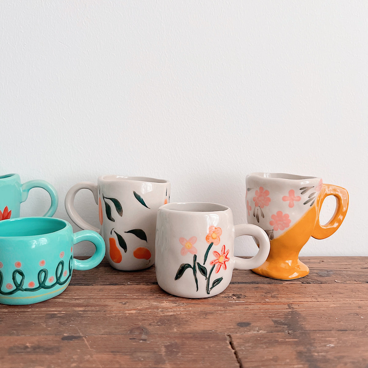 Close-up of a cream floral mug, a cream fruit-patterned mug, and a yellow-footed mug with pink flowers beside turquoise mugs.
