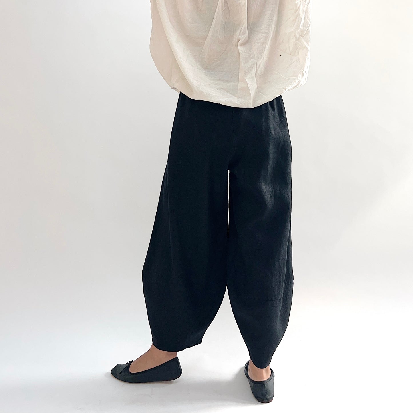 Back view of model wearing the Bryn Walker Heavy Linen Oliver Pant in Black, highlighting barrel-leg volume, elastic waistband, and tapered ankle in black heavy linen, styled with cream linen top and black ballet flats
