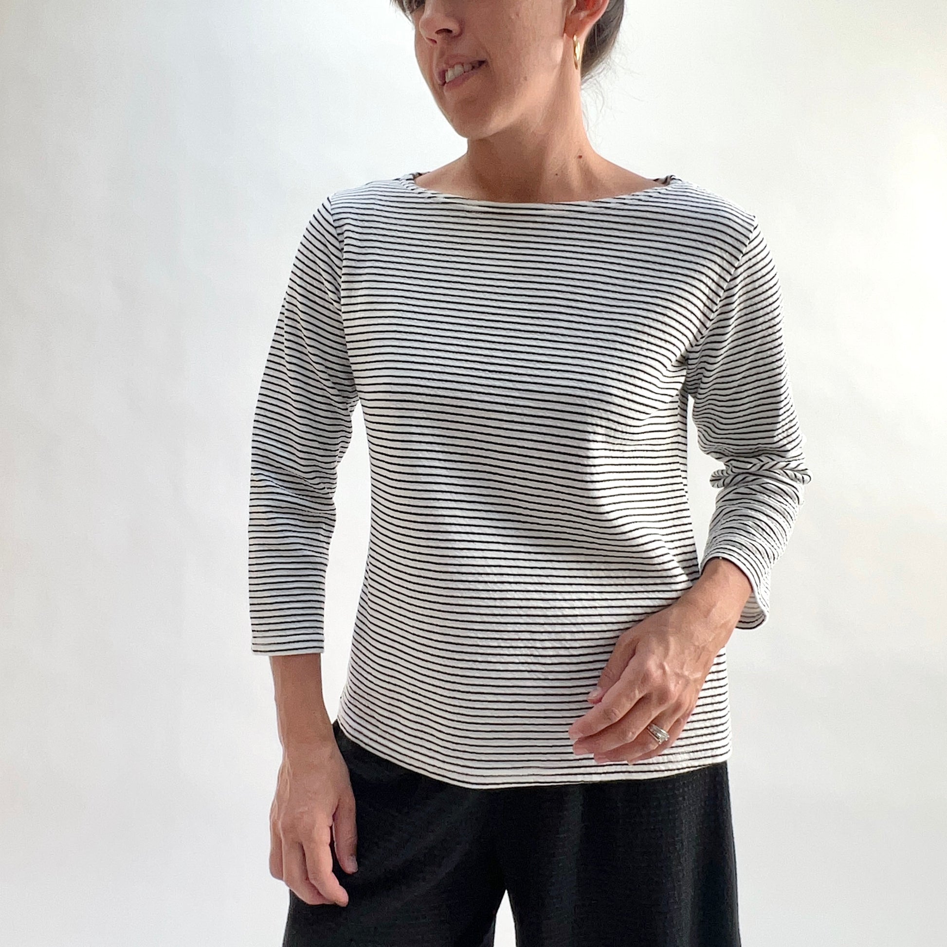 Full front view of model wearing a white and black horizontal stripe boatneck tee with 3/4 sleeves, hand at side, showing the relaxed fit, wide boatneck, and laundered crinkle-texture stripe, styled with black wide-leg pants against a white background.
