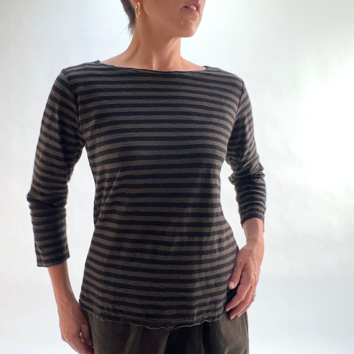 Front view of model wearing a chocolate and black horizontal stripe boatneck tee with 3/4 sleeves, hands at sides, showing the wide boatneck and relaxed silhouette, styled with dark olive pants against a white background.
