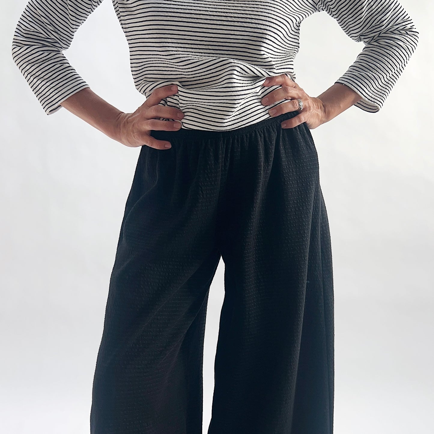 Close front view of model wearing black crinkle-texture cropped wide-leg pants, hands on hips at elastic waist, showing the fine crinkle-texture fabric and wide relaxed silhouette, styled with a white and black stripe long-sleeve top against a white background.
