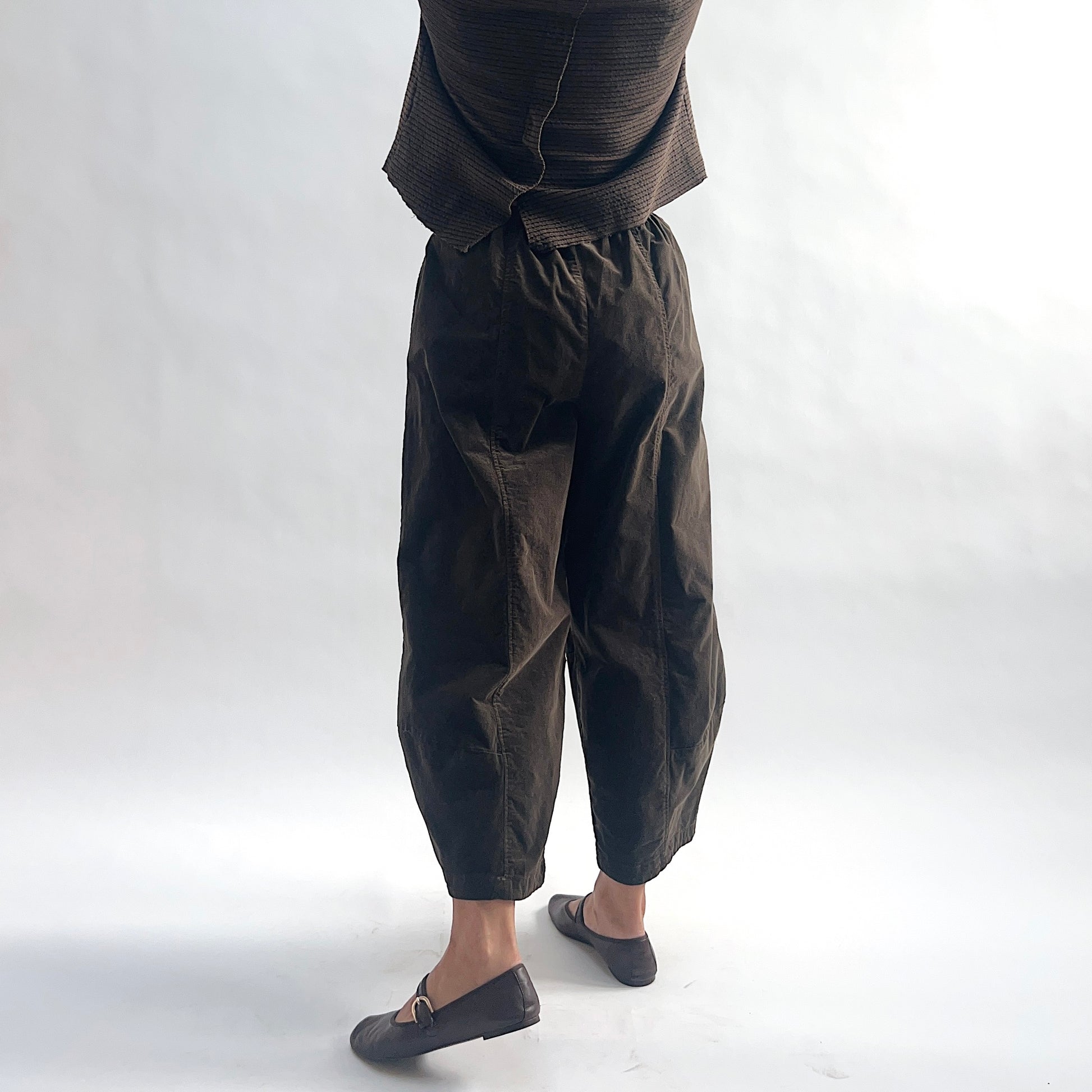 Three-quarter back view of model wearing chocolate mini cord lantern pants, showing the elastic waist gathering, rounded hip volume, tapered leg, and fine cord texture, styled with the matching striped cardigan and dark mary jane flats against a white background.
