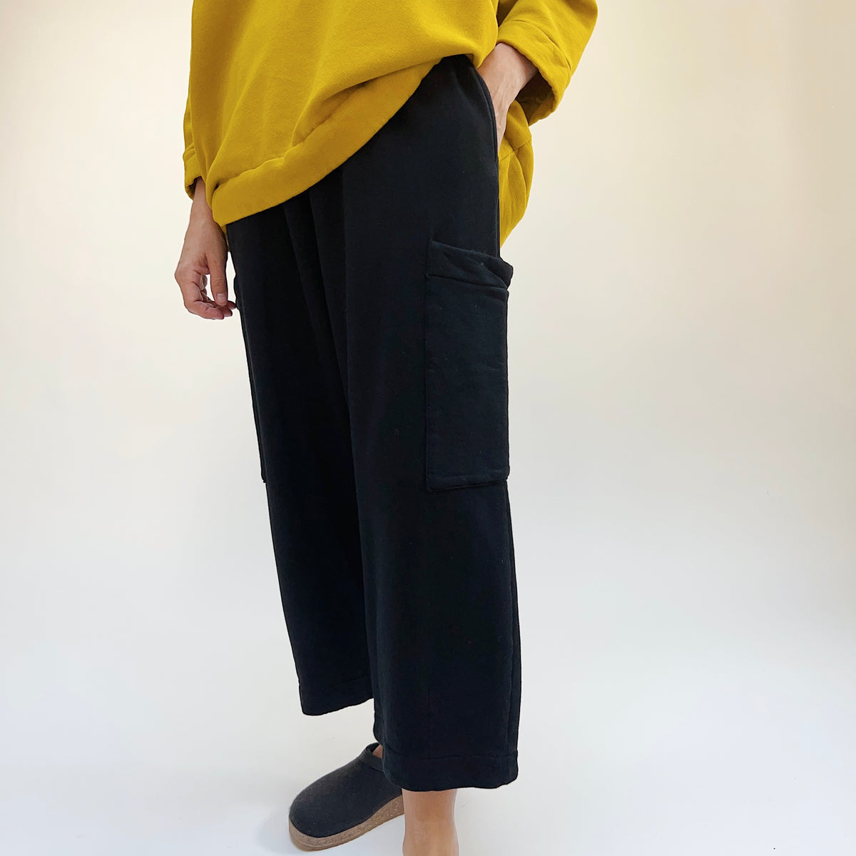 Three-quarter side view of a woman wearing the Bryn Walker Pasha Fleece Pant in Black, showing the elastic waistband, large thigh patch pocket, wide straight leg, and folded ankle hem in black organic cotton and bamboo fleece, styled with a mustard yellow sweatshirt and dark slide clogs against a white background
