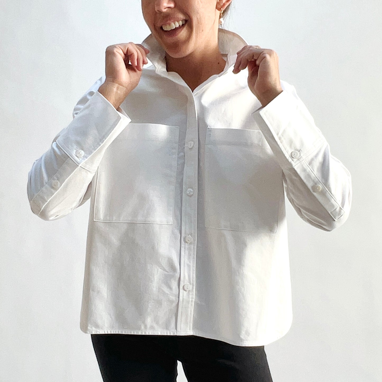 Close front view of smiling model wearing a white cotton oxford boyfriend shirt, hands raised to collar, showing the two large chest patch pockets, full button front, and wide barrel cuffs, styled with black pants against a white background.
