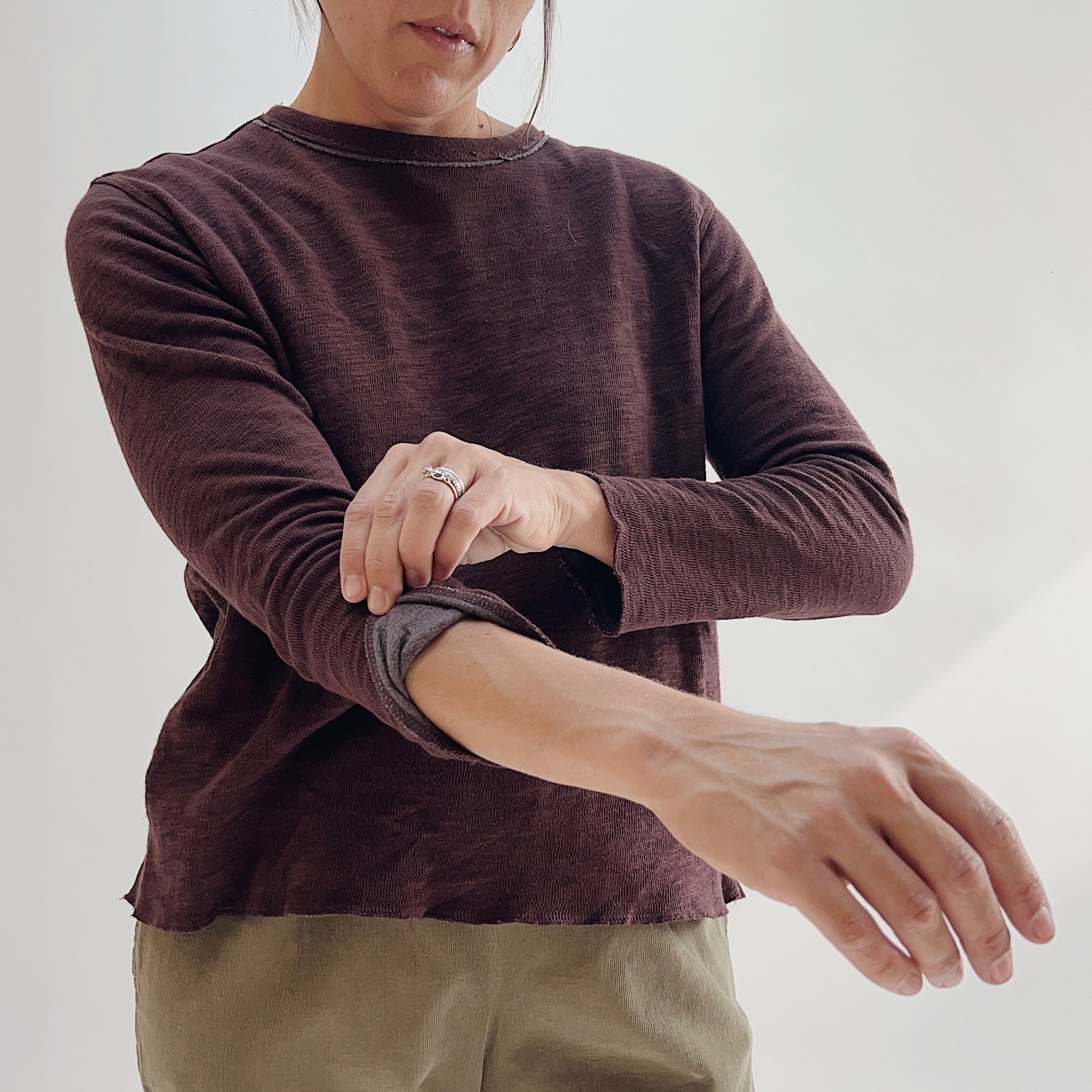 Close-up detail of the Bryn Walker Double Cotton Tee in Chicory, showing model rolling back sleeve cuff to reveal double-layer construction, raw-edge neckline, and textured chicory burgundy cotton blend fabric
