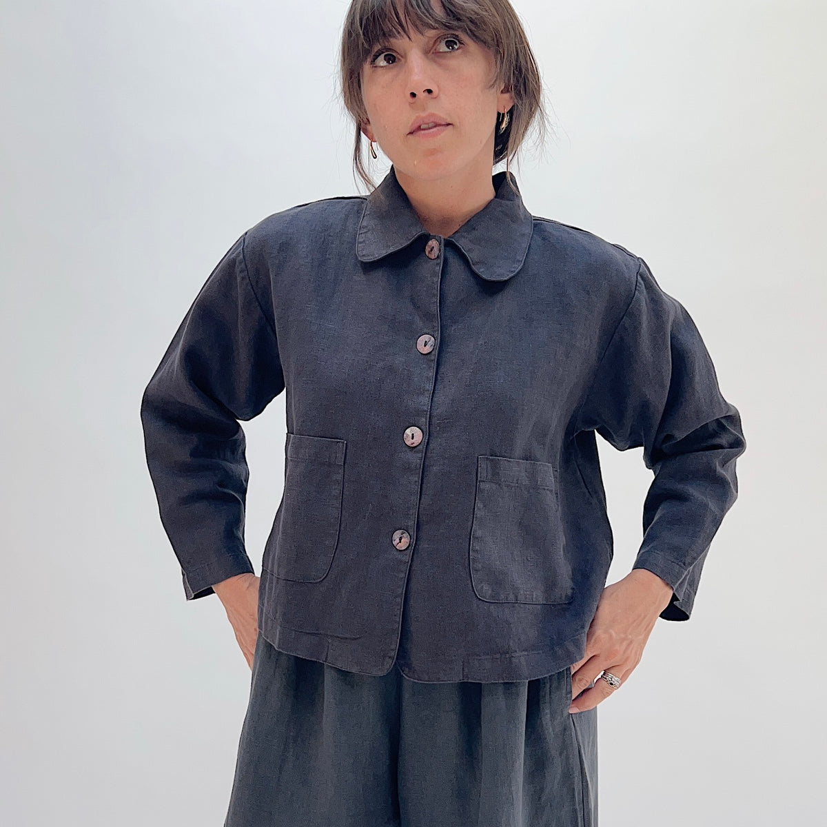 Model wearing the Bryn Walker Heavy Linen Audrey Jacket in Fig with hands on hips, showcasing relaxed cropped fit and shell buttons
